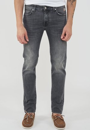 Uomo che indossa jeans neri sbiaditi slim-fit e scarpe da barca marroni, in piedi davanti a uno sfondo chiaro e semplice.