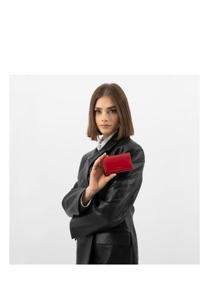 Jonge vrouw in een zwarte leren blazer die een kleine rode leren portemonnee vasthoudt, poserend tegen een effen witte achtergrond.