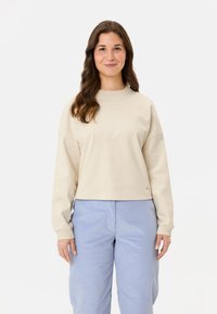 Beige verkürzter Sweatshirt mit langen Ärmeln, rundem Ausschnitt, weichem Textil und dezenten Akzenten am Saum, kombiniert mit hellblauen weiten Hosen.