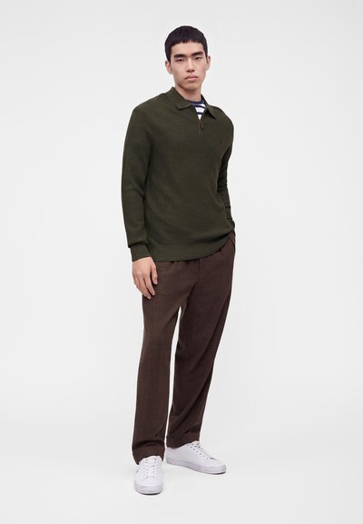 Homme debout portant un pull texturé vert olive, un pantalon marron à carreaux, des baskets blanches et une chemise rayée sous le pull.