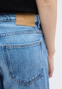 Πίσω τσέπη ανοιχτό μπλε τζιν με καφέ δερμάτινη ετικέτα που γράφει "τέλεια τζιν", φορεμένο από άτομο με μαύρο τοπ.