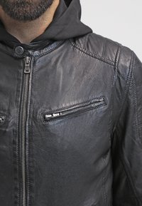 Schwarze Lederjacke mit Reißverschluss-Front, versehen mit vertikalen Reißverschlusstaschen und gesteppter Schulterverzierung. Enthält einen Kapuzenpullover darunter.