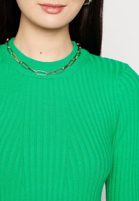 Top a maglia costina verde con scollo rotondo, caratterizzato da una collana d'oro con maglie ovali, che mette in risalto una finitura liscia e strutturata.