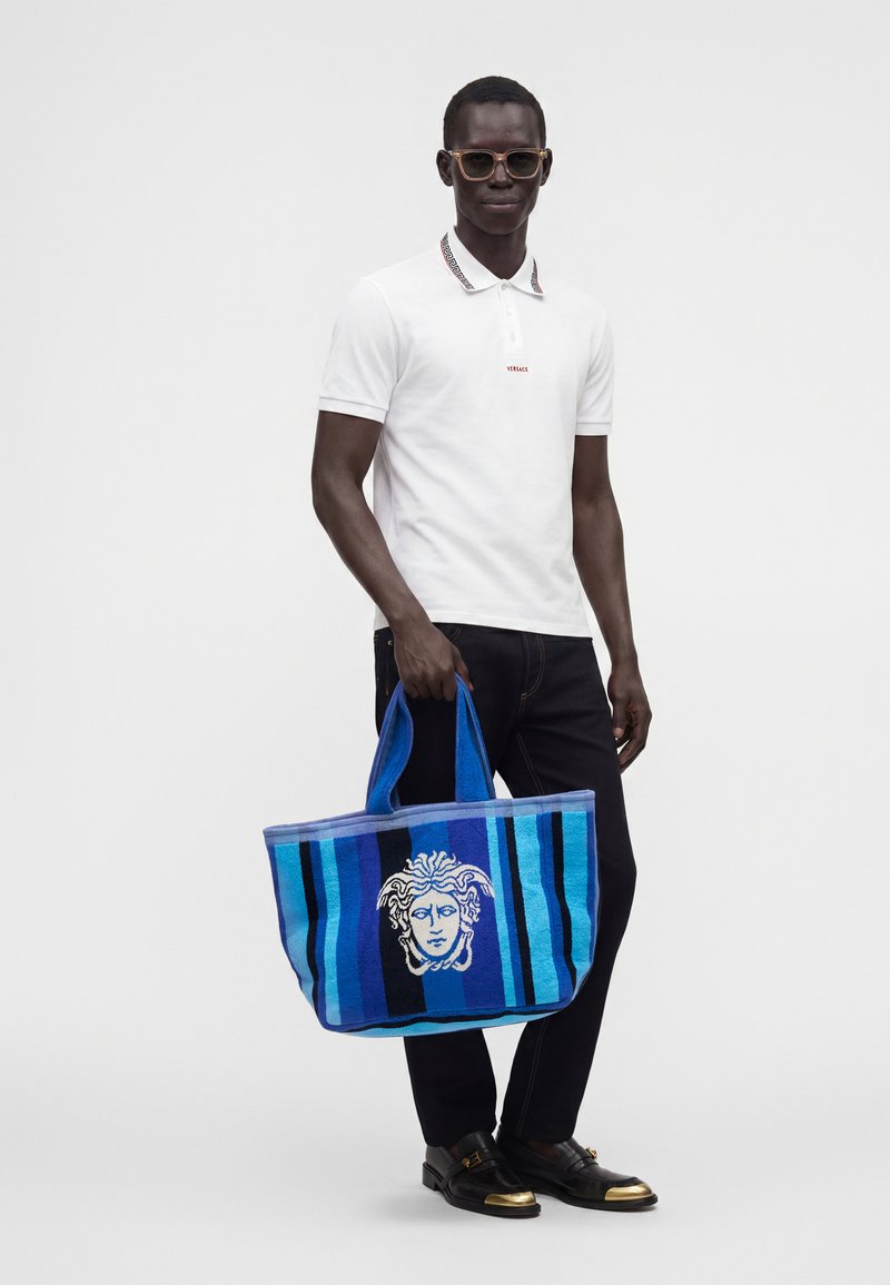 Homme portant un polo blanc, un pantalon noir, des lunettes de soleil et des chaussures noires, tenant un grand sac cabas bleu rayé avec un motif de tête de Méduse.
