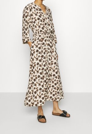 Mujer con un vestido largo blanco con estampado floral marrón, mangas tres cuartos, cintura ajustable con lazo y sandalias negras con tiras anchas con hebilla.