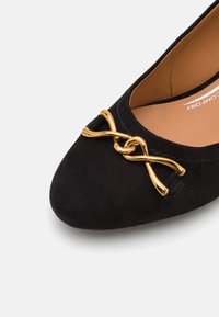 Ballerines en daim noir avec un bout rond et un accent en métal doré proéminent en forme de nœud à l'avant. L'intérieur est en cuir lisse.