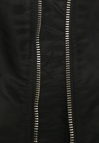Fermeture éclair métallique sur tissu noir avec un logo brodé subtil présentant un "A" stylisé et trois lignes horizontales de chaque côté.