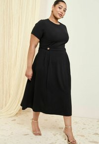 Schwarzes knielanges Kleid mit kurzen Ärmeln, das über einen taillierten Oberteil, ein Taillenband mit einem dekorativen Knopf und einen schwingenden Rock verfügt.