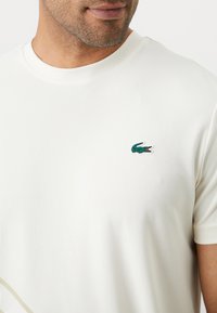 Camiseta de manga corta, color crema, hecha de un tejido texturizado, con el logo del cocodrilo verde de Lacoste en el lado izquierdo del pecho.