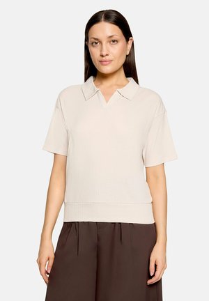 Femme aux cheveux lisses et foncés portant un haut beige à manches courtes avec col et un pantalon ample marron foncé sur un fond uni.