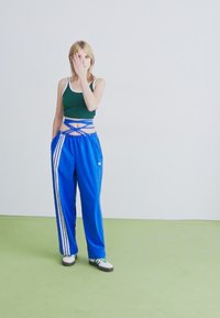 Fiatal nő zöld crop topban és élénk kék tréningnadrágban, amelyek keresztezett derékkal készült kötelekkel rendelkeznek, zöld padlón állva, egyik szemét a kezével eltakarva.