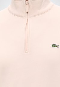 Pull rose clair à zip quart, col montant et logo crocodile vert brodé sur la poitrine gauche.