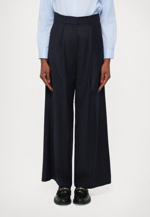 Calças largas em azul escuro, com frente pregueada, cintura ajustada e um tecido liso e texturizado. Combinadas com sapatos pretos.