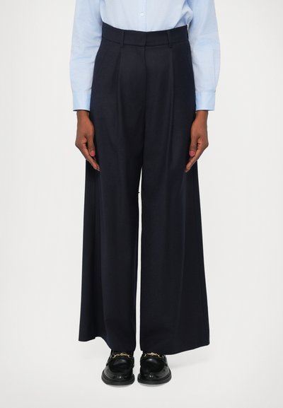 Pantalons larges en bleu marine foncé, avec un devant plis, une taille ajustée et un tissu lisse et texturé. Assortis à des chaussures noires.