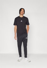 Melnā T-krekls ar baltu logo, kombinēts ar melniem sporta biksēm ar sānu kabatām un baltām akcentiem, valkāts ar baltiem sporta apaviem.