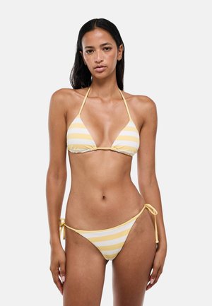 Femme portant un bikini halter rayé jaune et blanc avec des bas à nouer sur les côtés, debout devant un fond blanc uni.