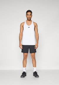 Débardeur de sport blanc sans manches avec logo noir, associé à un short foncé. Le mannequin porte des chaussettes blanches et des chaussures de sport noires. Fond neutre.