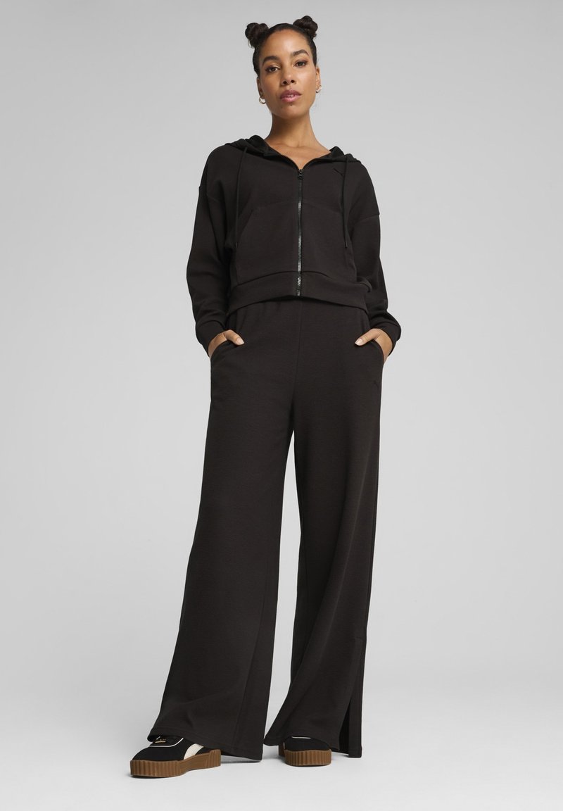 Black zip-up cropped hoodie with a front pocket, paired with wide-leg black pants. Smooth fabric with a relaxed fit. Black platform sneakers.