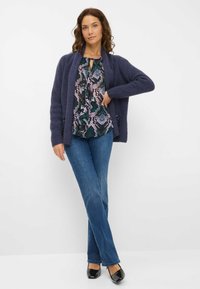 Marineblå strikket cardigan lagt over en mønstret bluse, parret med blå jeans og sorte lakerede sko. Har åben front og tekstureret stof.