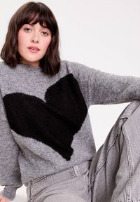 Grå tekstureret sweater med et stort sort hjerteprint foran, brede ærmer og en afslappet pasform. Sammen med grå jeans.