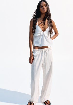 White linen crop top with a v-neck and ruffled detail, paired with loose, cream-colored pants and black flip-flops on a light background.