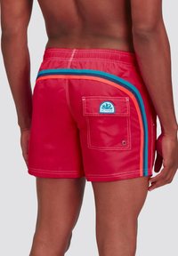Costume da bagno rosso realizzato in materiale lucido, con strisce blu e arancioni, una tasca posteriore con un patch logo e cuciture bianche in evidenza.