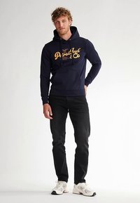 Tmavě modrá mikina s oranžovým nápisem, kapucí na šňůrku a klokankovou kapsou, v kombinaci s černými džínami a bílými teniskami.