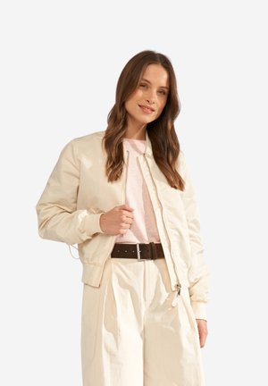 Žena s dlhými hnedými vlasmi, ktorá nosí krémovú bomber bundu, svetloružové tričko, krémové nohavice s vysokým pásom a tmavohnedý opasok, mierne sa usmievajúca.