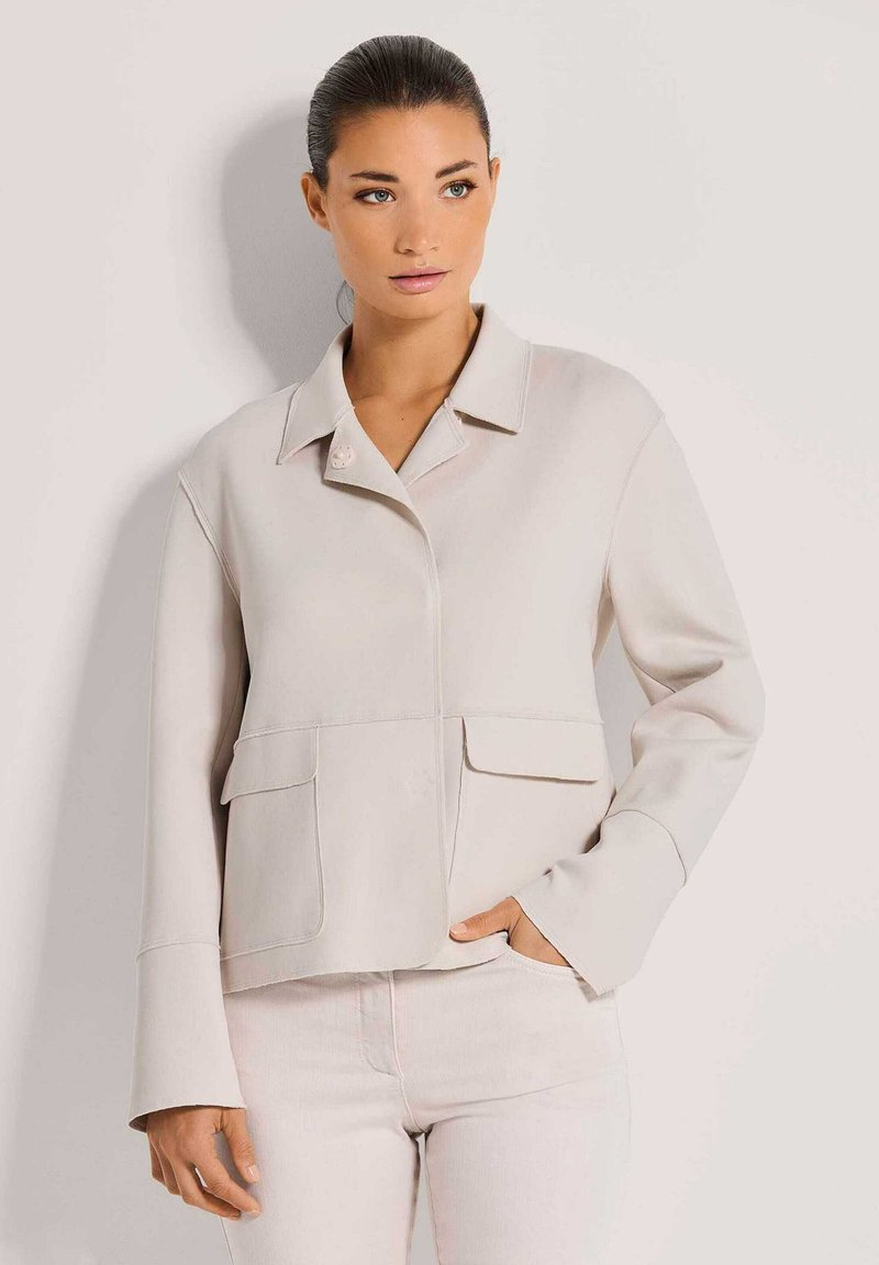 Frau mit zurückgegeltem Haar trägt eine hellbeige Jacke mit Klappentaschen und passende helle Hose, steht vor einer schlichten Wand.