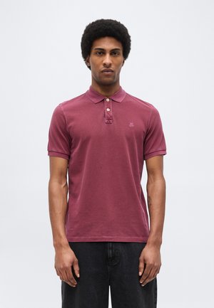 Rövid, göndör hajú férfi, aki egy kifakult bordó rövid ujjú galléros pólót és fekete nadrágot visel, egyszerű háttér előtt áll.