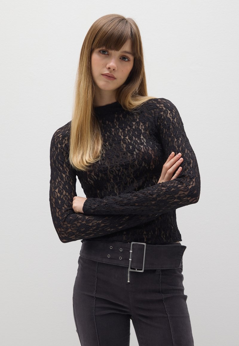 Haut en dentelle noire à manches longues avec un col montant, présentant un motif floral. Associé à un pantalon sombre taille haute et une ceinture avec boucle rectangulaire.
