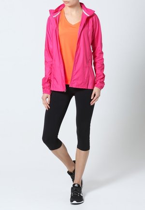 Giacca leggera rosa con cappuccio, t-shirt arancione sotto, leggings a capri neri e scarpe da ginnastica nere. Tessuto morbido e design aderente.
