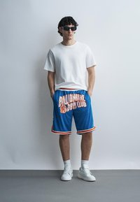 Moški, ki nosi črna sončna očala, belo majico, modre kratke hlače z napisom "Billionaire Boys Club", bele nogavice in bele superge, stoji ob običajni steni.