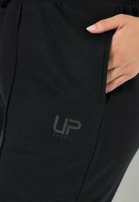 Černá mikina s měkkou texturou, která má boční kapsu a tištěnéLogo „UP SPORTS“ v šedé barvě na dolní části.