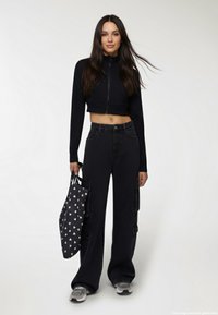 Czarna krótka kurtka zapinana na zamek, spodnie cargo w kolorze czarnym z wysokim stanem i bocznymi kieszeniami, trzymając ciemną torbę z małym białym wzorem serca oraz szare sneakersy.