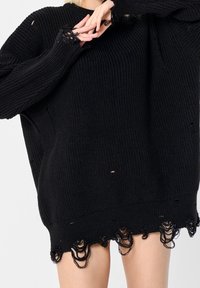 Personne portant un pull en tricot noir surdimensionné avec des bords usés et de petits trous, les mains jointes près de la poitrine, les jambes visibles sous l'ourlet.