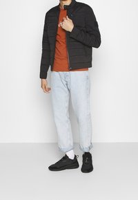 Black puffer jacket with a zipper and stand collar, worn over an orange t-shirt. Light blue tapered jeans and black sneakers complete the look.