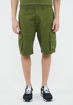Uomo con pantaloncini cargo verde oliva e sneakers nere Saucony Jazz, in piedi davanti a uno sfondo chiaro e semplice.
