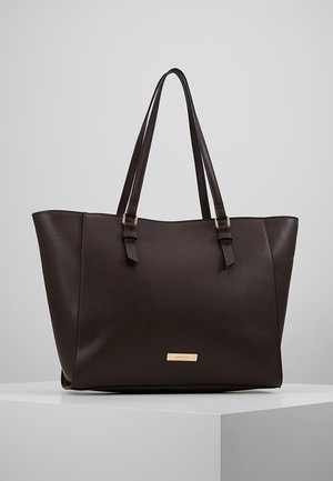 Mørkebrunt lædertotetask med to lange håndtag og en lille rektangulær guldfarvet mærkeplade foran, vist på et hvidt podium.