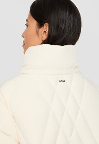 Woman with dark hair wearing a high-collar cream quilted jacket with "BOSS" logo on the back.