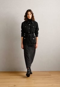 Completo in denim nero composto da una giacca con bottoni e vita legata, e una lunga gonna. Abbinato a stivali alla caviglia. Textura liscia nel complesso.