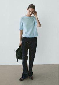 Maglione azzurro chiaro a maniche corte con scollo e orlo a coste, abbinato a pantaloni scuri a zampa e scarpe marroni. Design semplice, texture morbida.