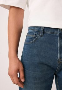 Sötétkék farmernadrág, amely testhezálló kialakítással rendelkezik, elülső zsebbel, övhurkokkal és látható varrási részletekkel. Az anyag kopott textúrájú.