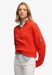 Pulls en laine torsadée, court, de couleur rouge vif avec un col zippé. Présente des poignets côtelés et un patch sur la manche gauche. Porté sur une chemise à rayures bleu clair.