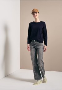 Ležérní outfit obsahující tmavě modrou košili s dlouhým rukávem a šedé volné džíny. Doplněný je hnědým kšiltem a světlými teniskami.