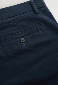 Granatowe chinosy o miękkiej teksturze, z tylną kieszenią oraz okrągłym guzikiem z czterema dziurkami nad kieszenią do zapięcia.