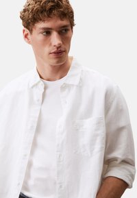 Camisa blanca de lino con botones, con un bolsillo en el pecho, mangas arremangadas, combinada con una camiseta blanca de cuello redondo debajo. Textura suave.