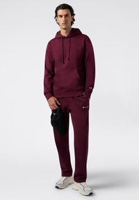Completo in morbido tessuto composto da felpa e pantaloni della tuta color bordeaux con accenti di logo. Il modello tiene in mano un cappellino nero; le sneakers sono di colore crema con dettagli testurizzati.