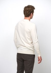 Maglione a maniche lunghe color crema con scollatura rotonda, morbida texture, polsini e orlo a costine; indossato con pantaloni marroni texturizzati.