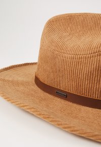 Brun cordfløyelsbredbremmet hatt med brunt lærbånd som har et metalllogoaccent. Teksturert finish med flat krone og flettet detalj.
